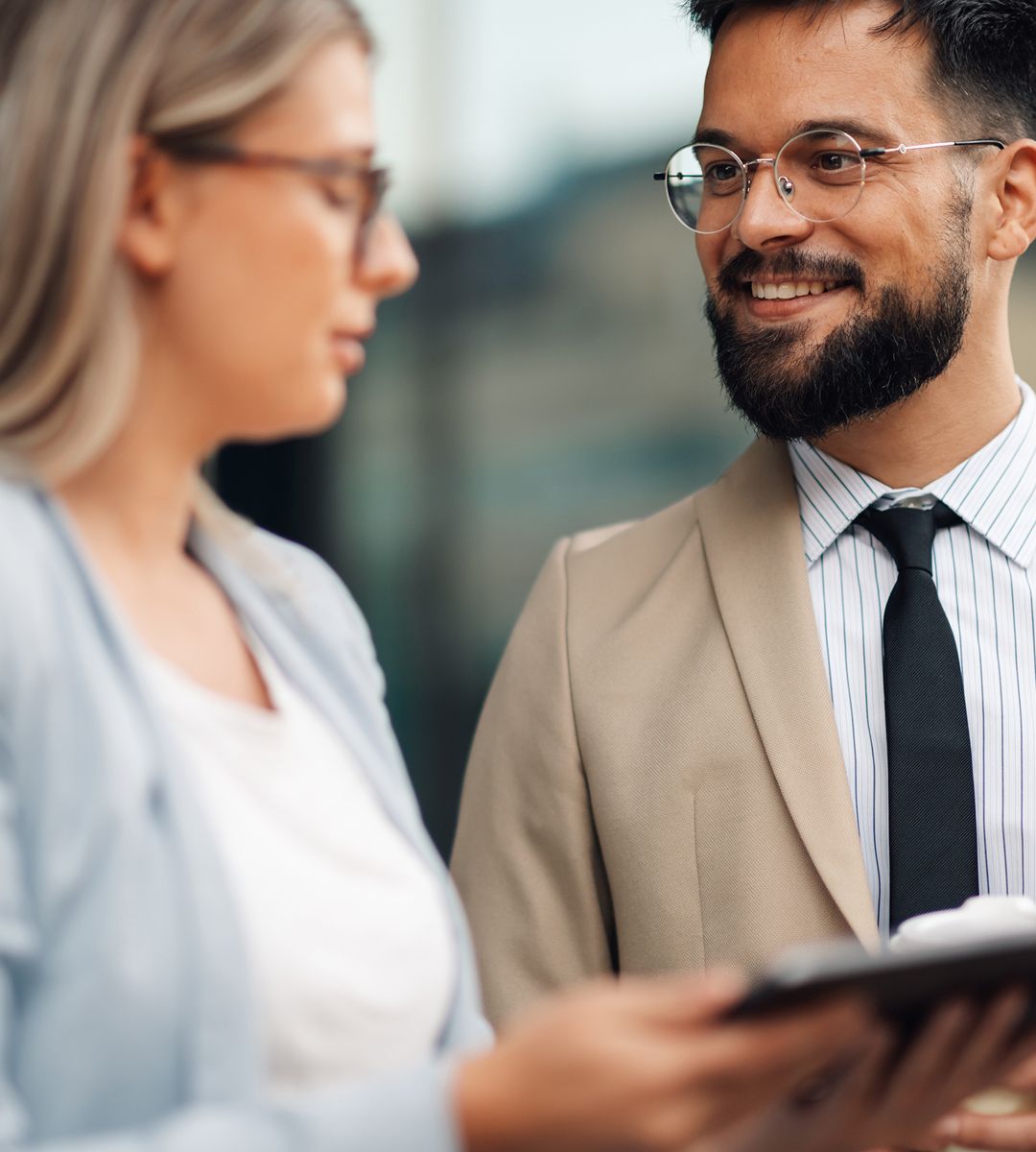 Two individuals engaged in a conversation outdoors, one holding a tablet, both dressed in professional attire, with happy expressions.