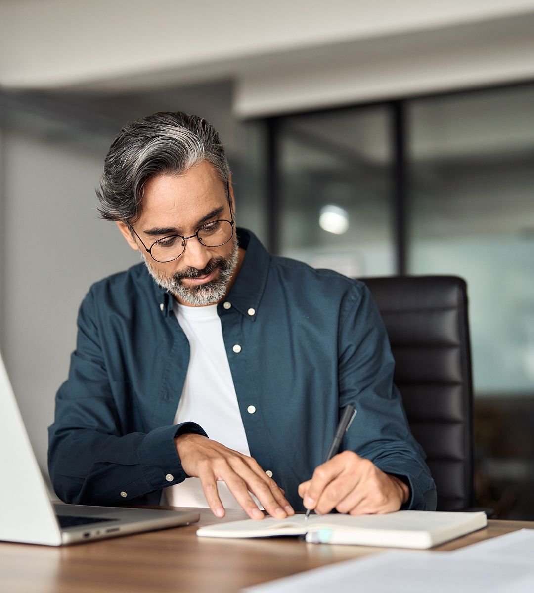 Person seated at a desk, writing in a notebook, with a laptop and modern office background. Features casual attire and glasses.