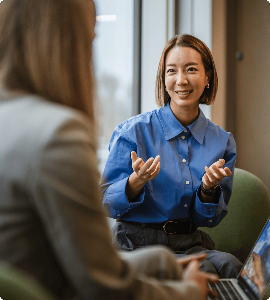 A person in a blue shirt gestures while speaking to another individual, seated together in a bright, modern setting.