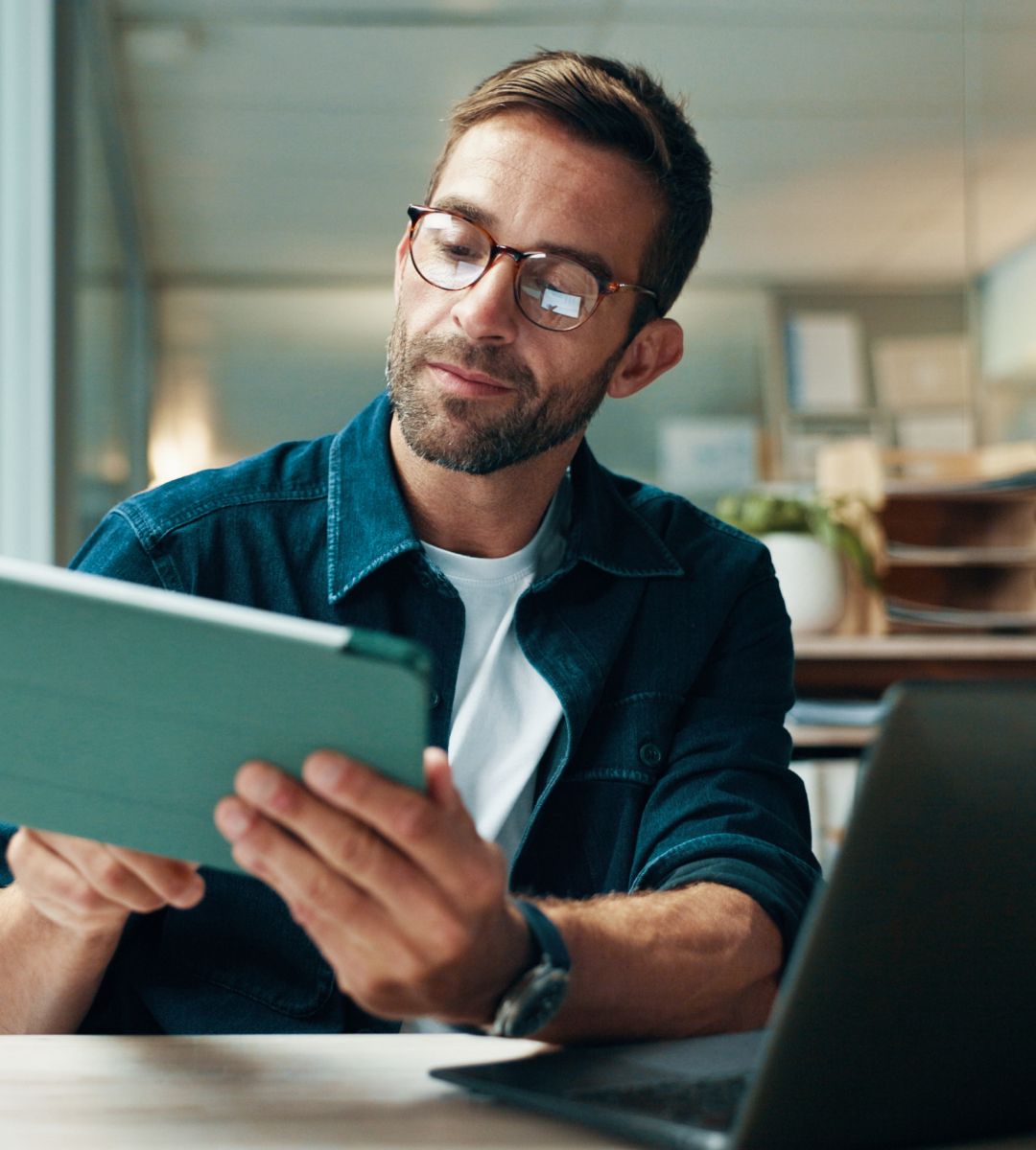A person reading a tablet in an office setting, with a laptop nearby and a casual, focused expression.
