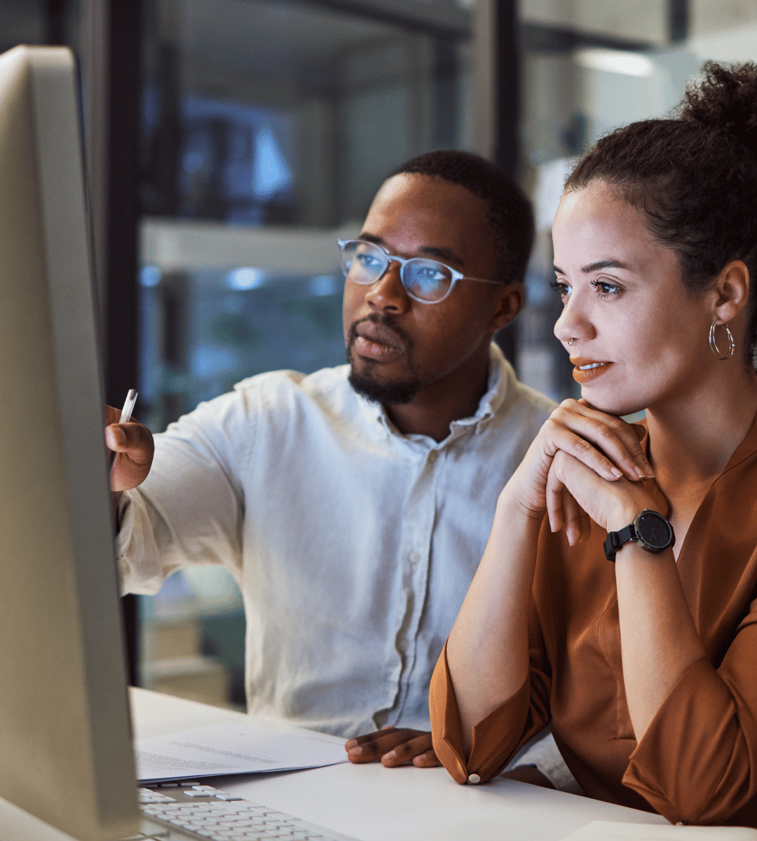 Two individuals collaborating at a computer, focusing on a screen in a modern office environment, engaged in discussion.
