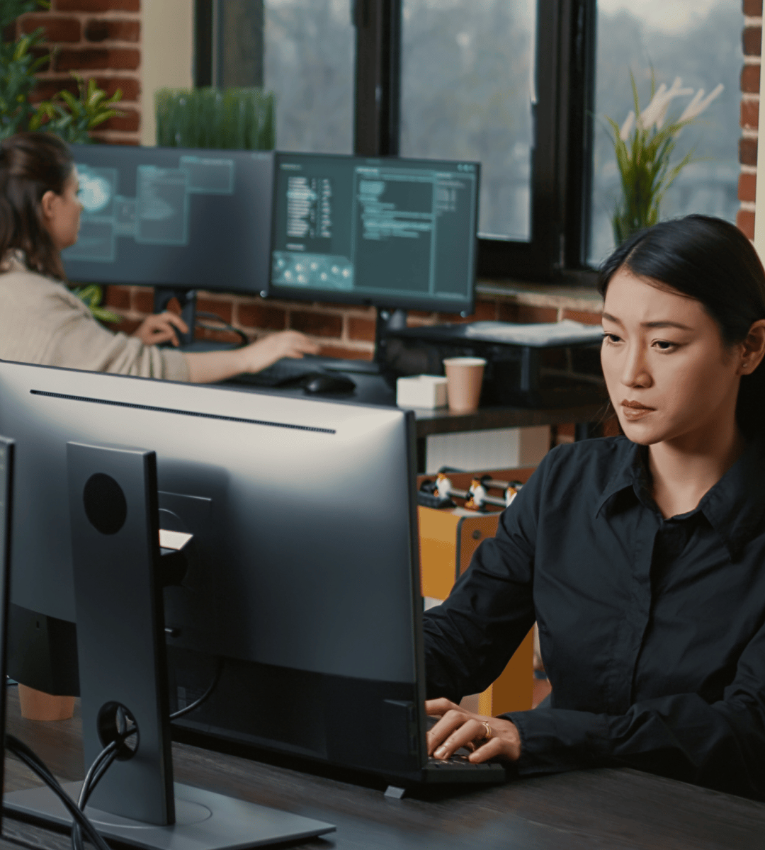Two individuals work at computers in a modern office environment, focused on their tasks with multiple monitors visible.
