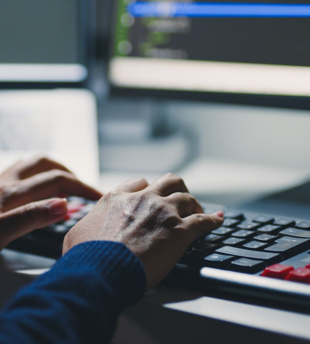 Hands typing on a keyboard in front of a computer screen displaying coding or programming content.