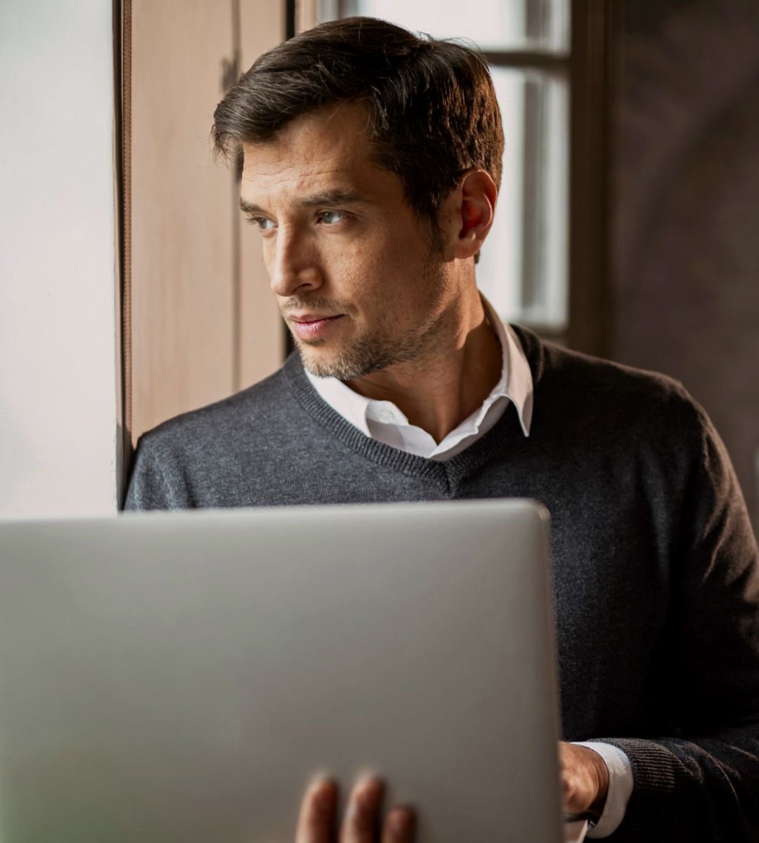 Professional headshot of a person using a laptop while looking thoughtfully out a window, wearing a dark sweater over a collared shirt.