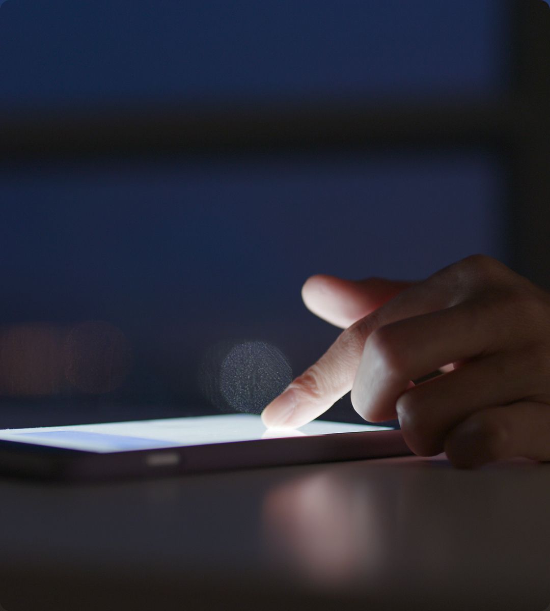 Close-up of a hand tapping a tablet screen in low light, with a blurred background and soft glow from the screen.