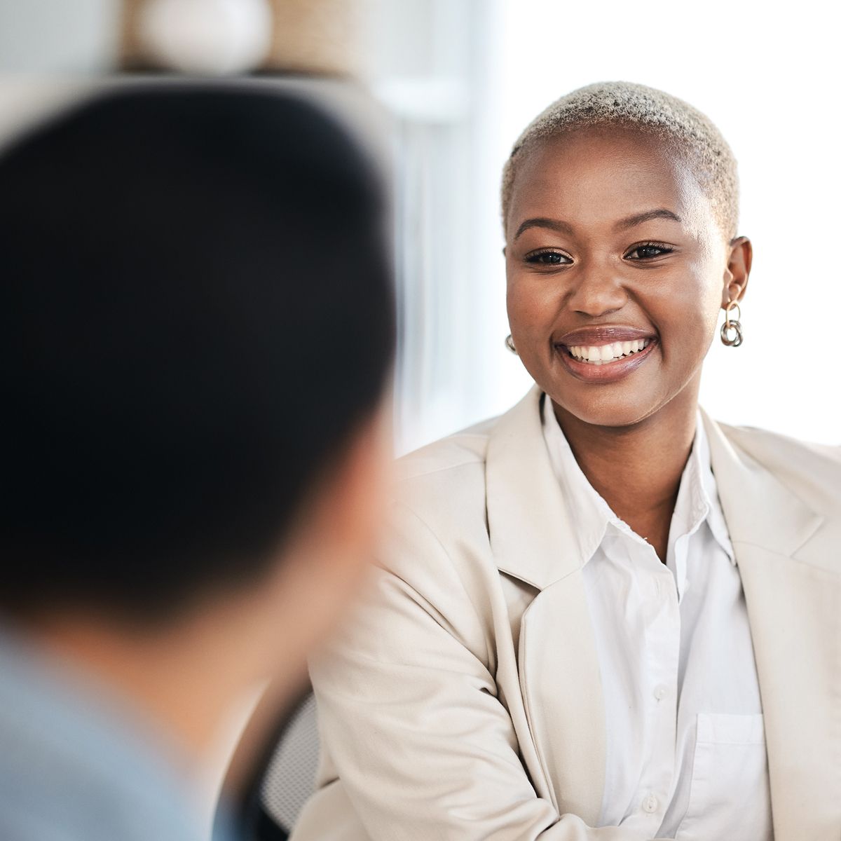 Smiling individual with short hair wearing a light blazer and earrings, engaged in conversation with another person out of focus.