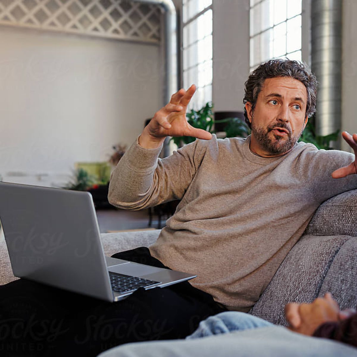 A person engaged in conversation while gesturing, sitting on a couch with a laptop open beside them in a bright interior space.