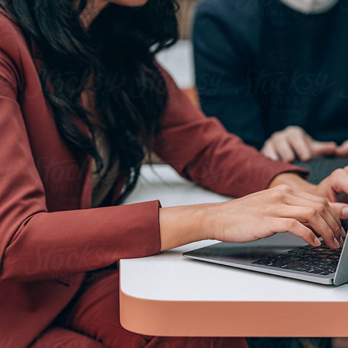 Hands typing on a laptop at a table, with another person visible in the background, suggesting a collaborative work environment.