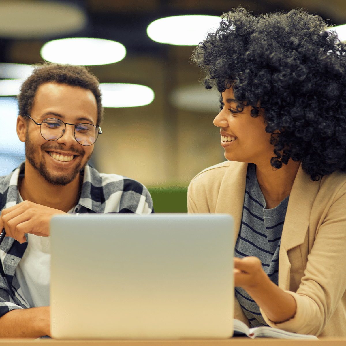 Two individuals engaged in a discussion while working on a laptop in a casual workspace. The atmosphere is collaborative and friendly.