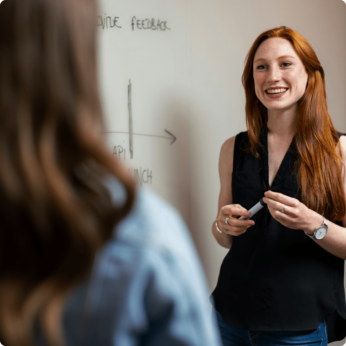 Person with long red hair smiles while holding a marker, standing in front of a whiteboard with feedback notes.