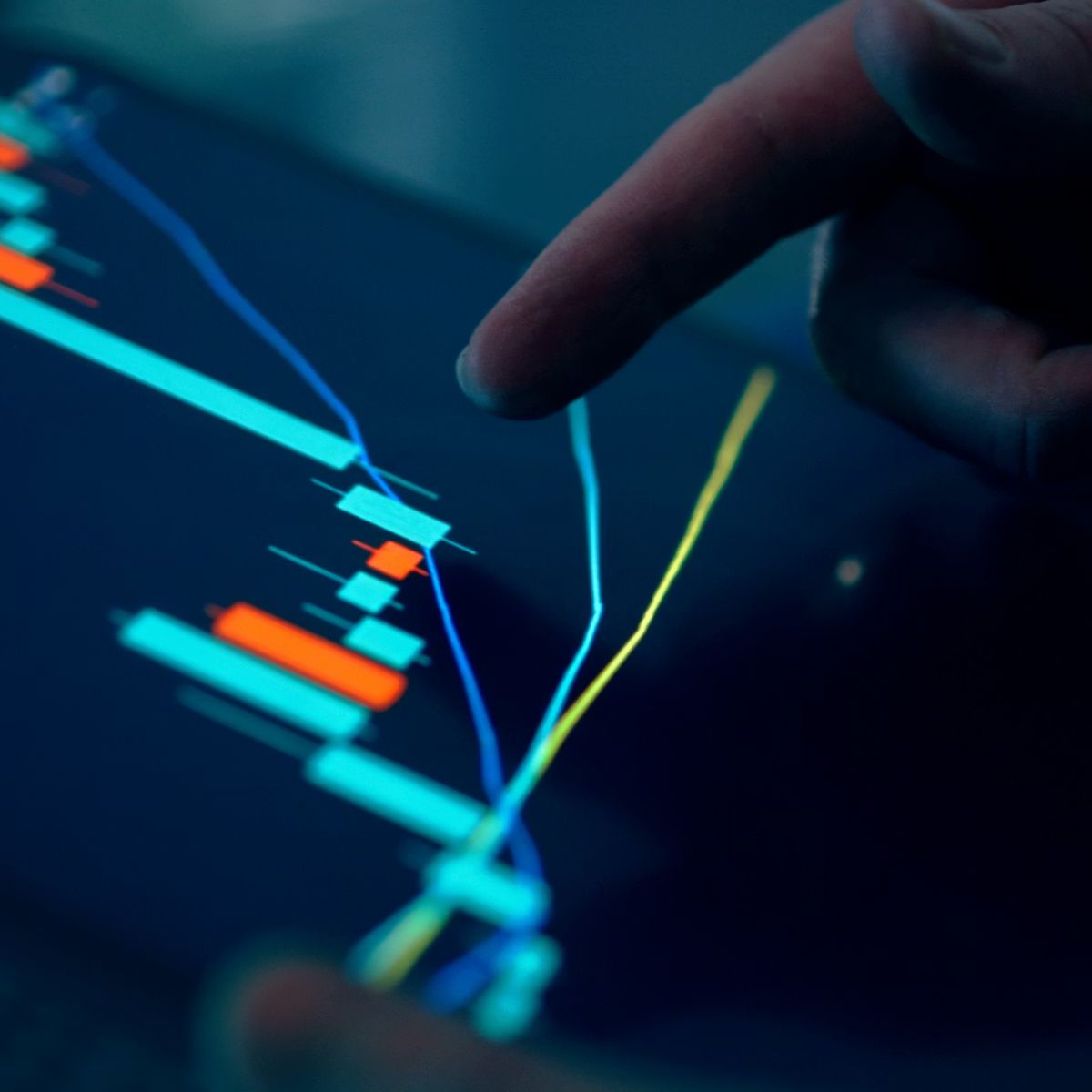 A close-up of a hand pointing at a digital screen displaying a colorful financial chart with various lines and bars.