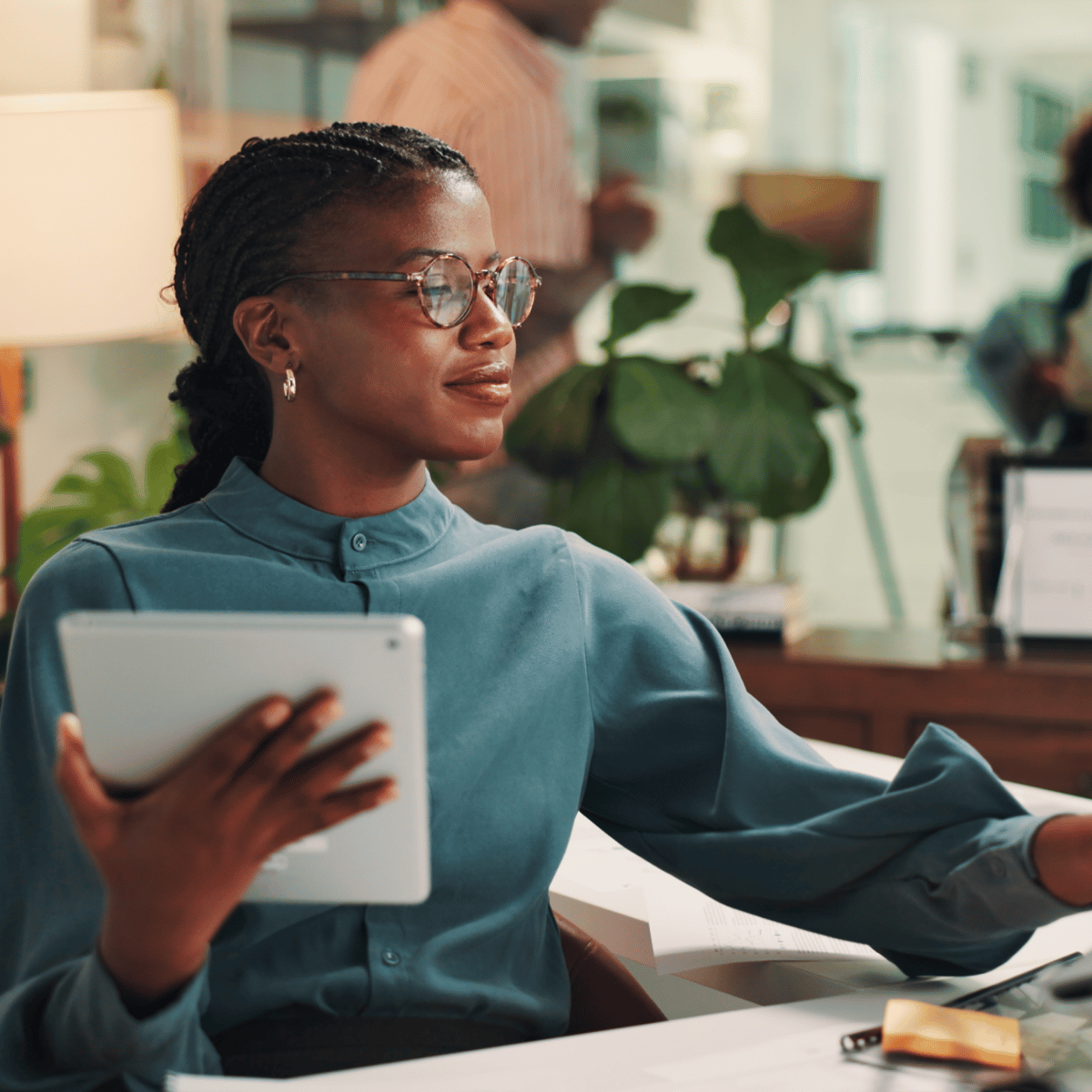 Professional in an office setting, holding a tablet and engaging with a computer, surrounded by plants and a collaborative workspace.