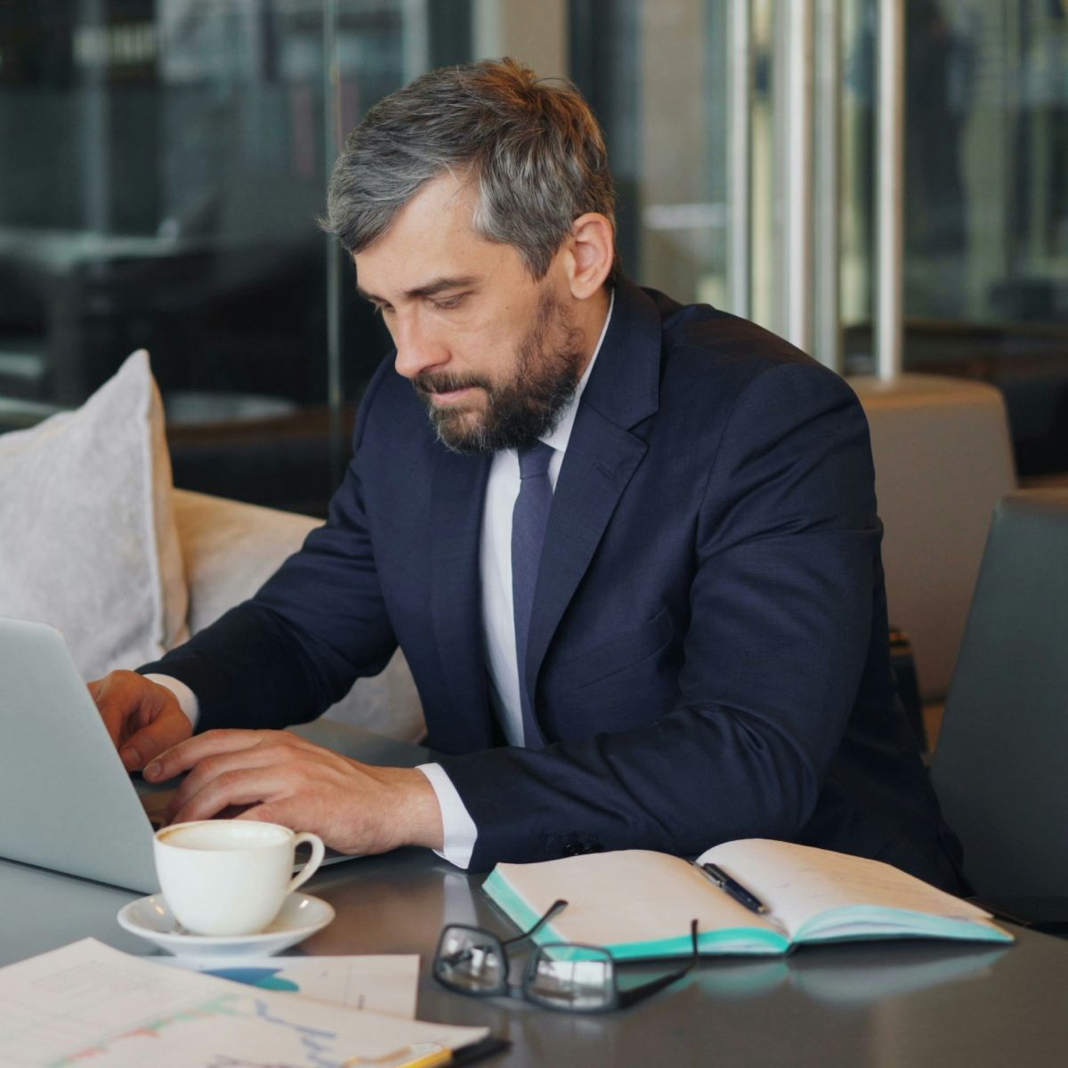 A professional sitting at a table working on a laptop with a cup of coffee and an open notebook nearby.