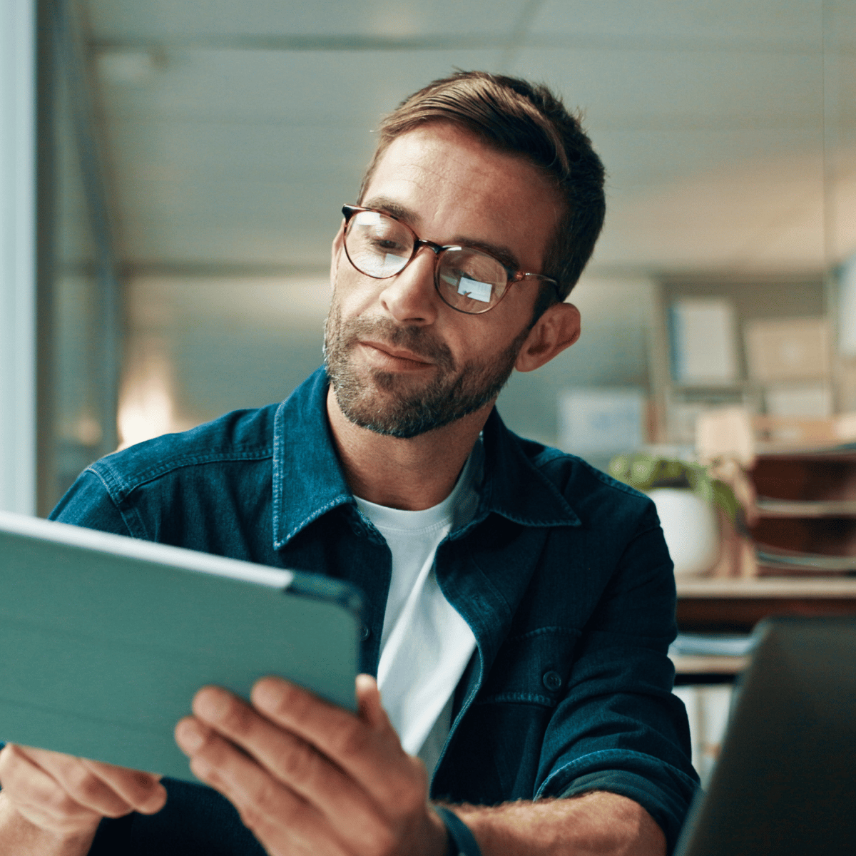 A person wearing glasses is focused on a tablet while seated in an office environment with natural lighting and a tidy workspace.