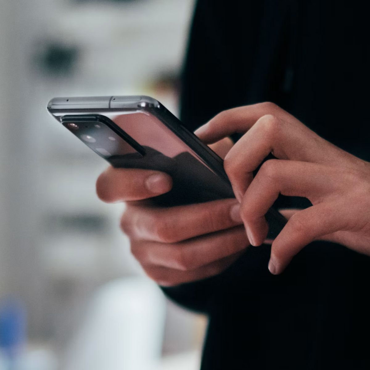 A person using a smartphone, focusing on the device with both hands, in a blurred indoor setting.