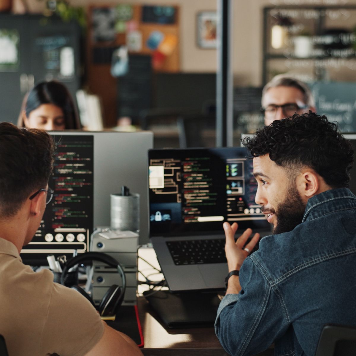 A group of individuals engaged in a discussion about coding and technology at a modern workspace. Several laptops and screens display code.