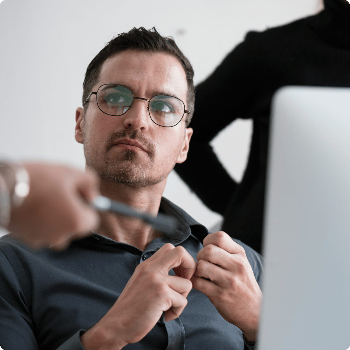 Person sitting at a desk, looking intently at a computer screen, with hands positioned on shirt, another person standing nearby.