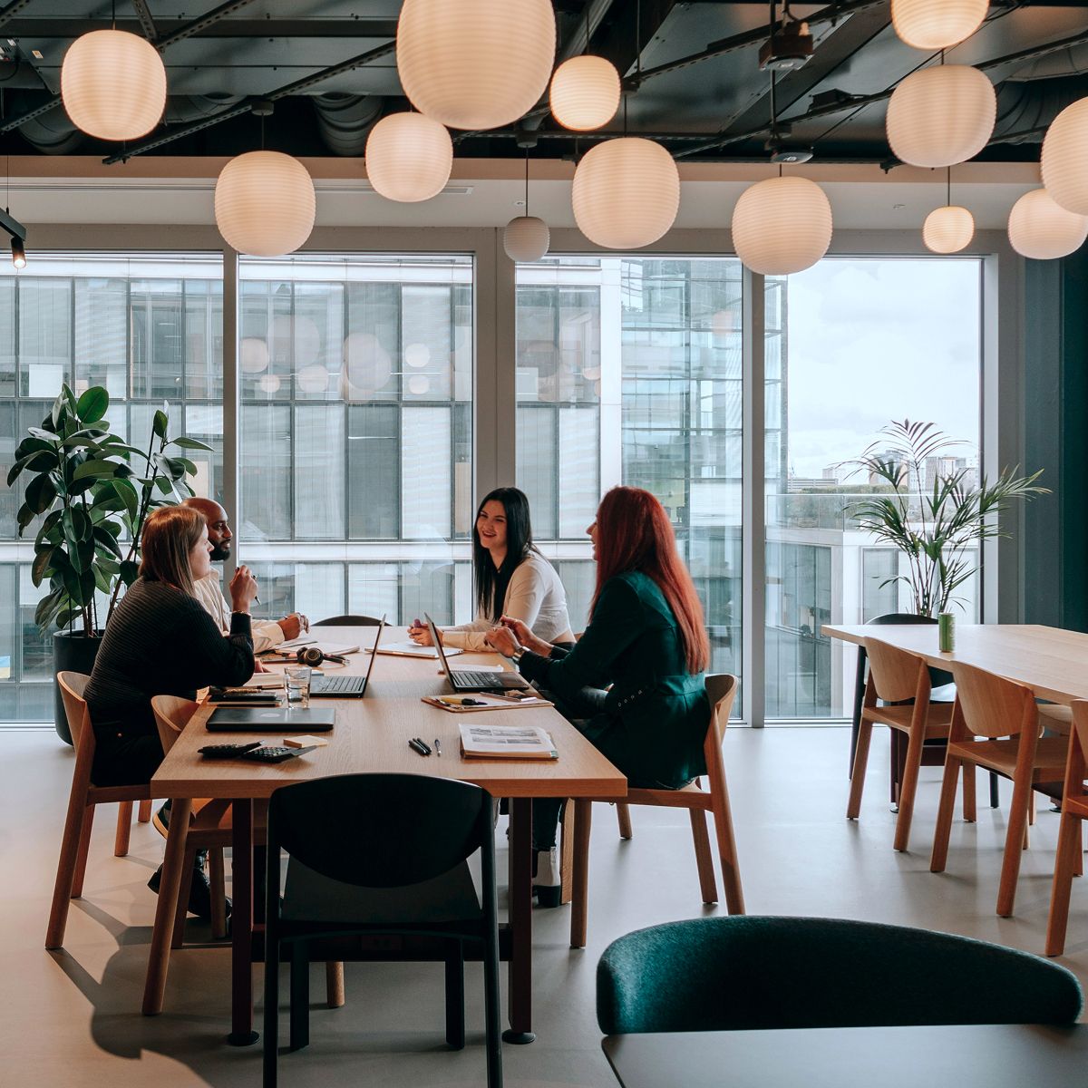 A group of people engaged in discussion around a conference table in a modern office with large windows and pendant lighting.