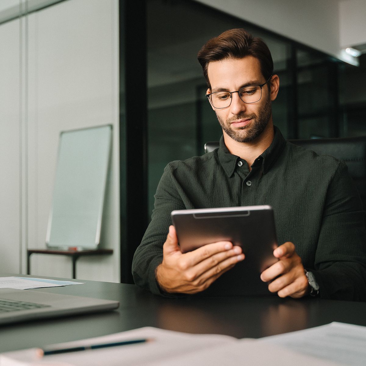 Person sitting at a desk holding a tablet, wearing glasses and a dark shirt, with a laptop and documents visible on the table.