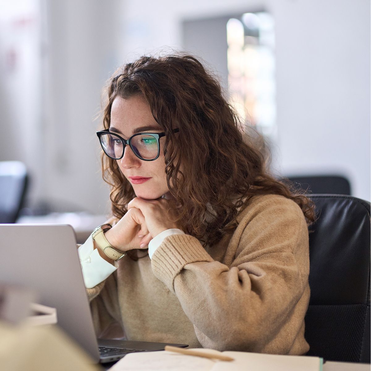 Person with curly hair and glasses focused on a laptop, seated at a desk in a bright, modern office environment.