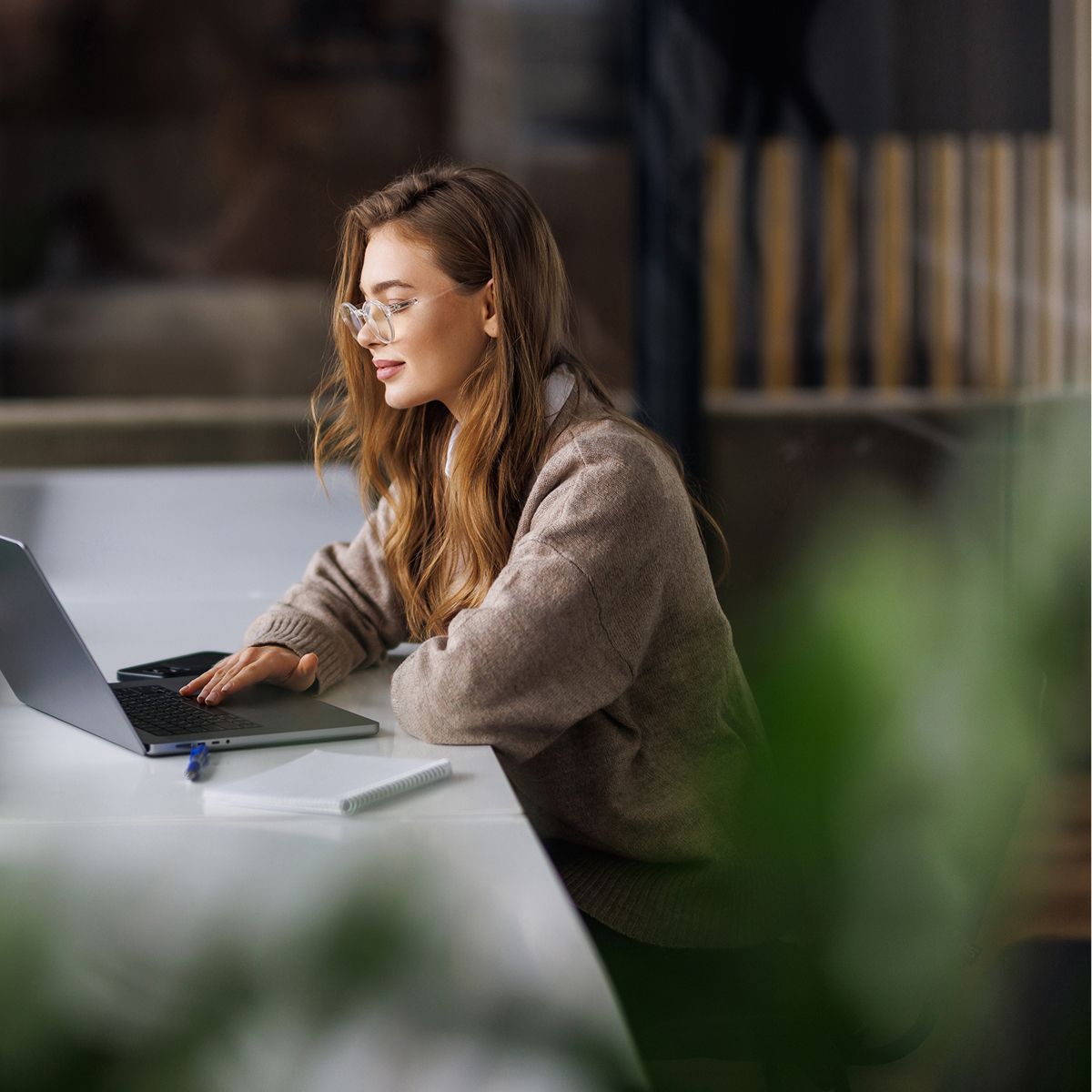 A person with long, wavy hair sits at a table, working on a laptop, wearing glasses, surrounded by greenery in a modern workspace.