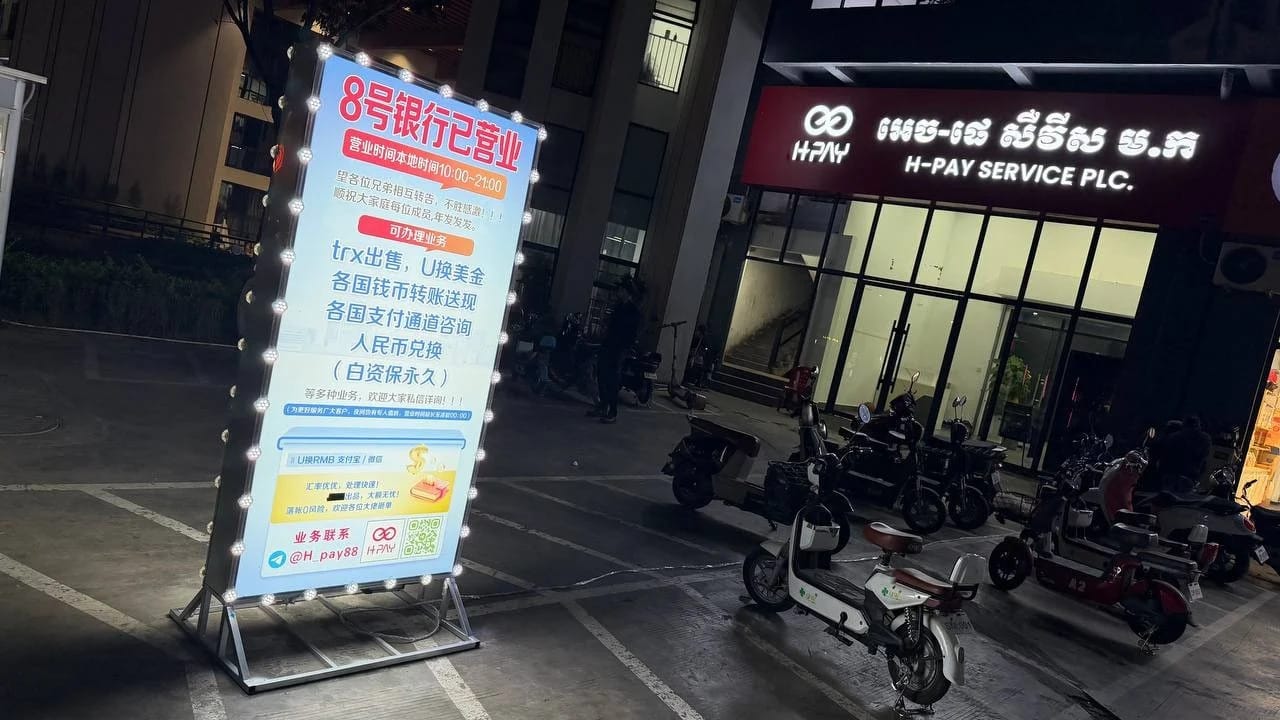 A lit sign displaying advertisements and information outside a service building, surrounded by parked scooters and dimly lit surroundings.