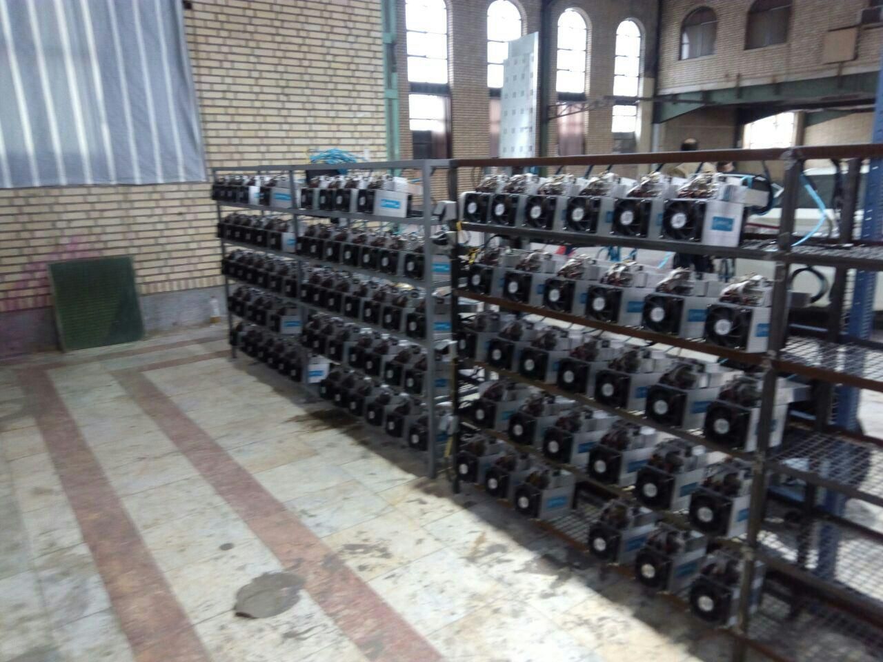 Rows of cryptocurrency mining equipment in an industrial setting with brick walls and large windows.