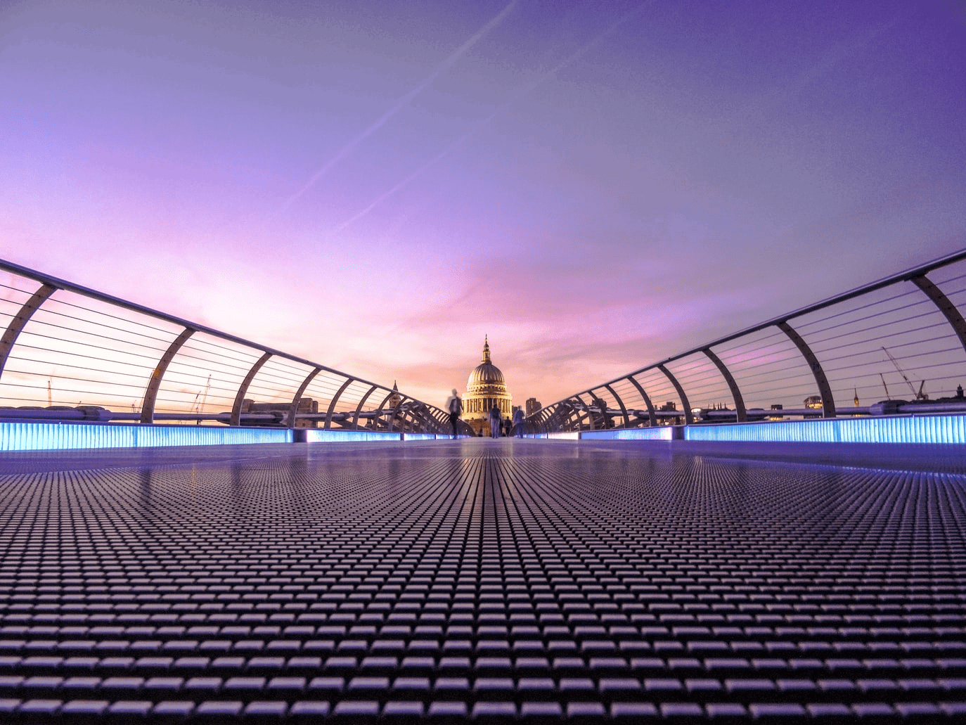 View of a modern bridge leading towards a dome structure at sunset, with a vibrant purple sky and illuminated path.