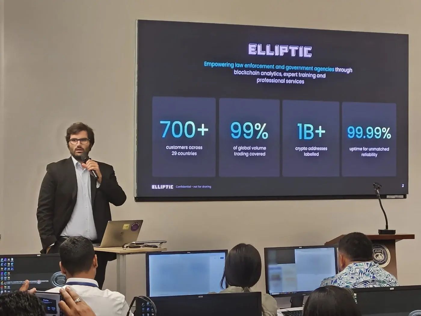Presentation in a professional setting with a speaker discussing blockchain analytics, supported by visuals on a screen, while audience members listen.