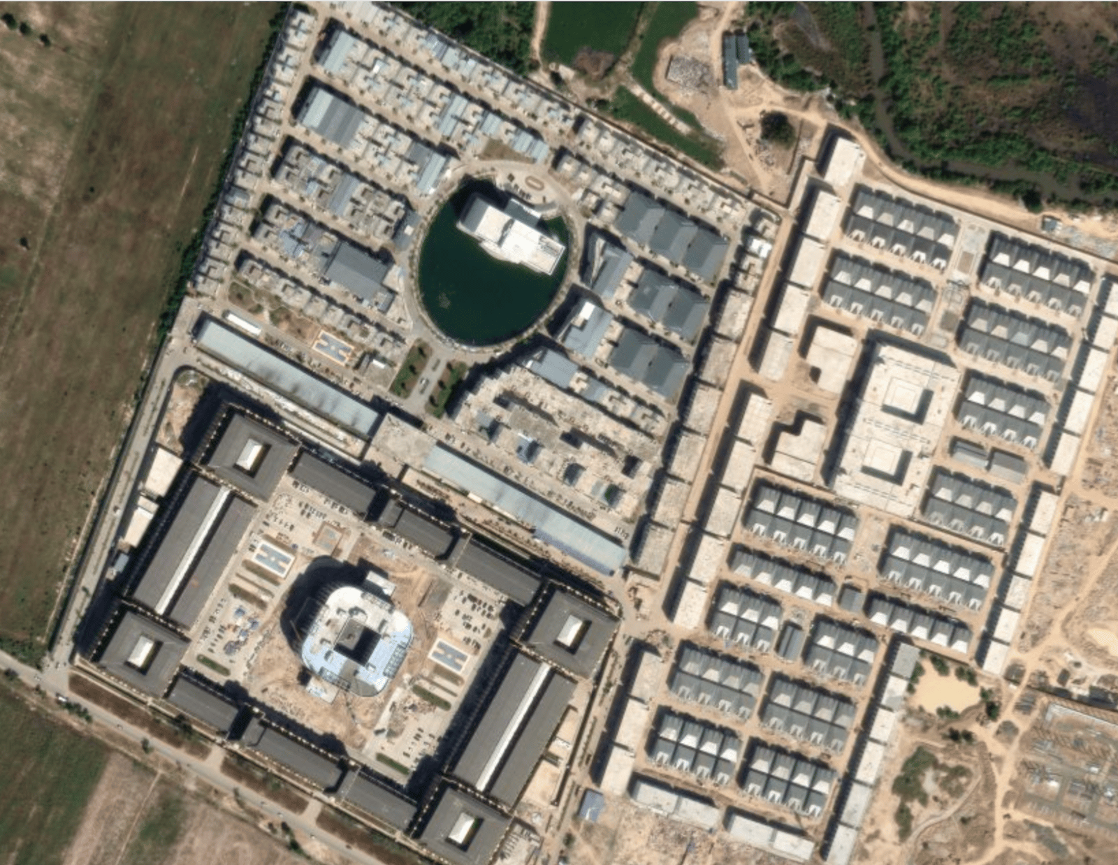 Aerial view of a large facility with multiple buildings arranged in geometric patterns, surrounded by greenery and a body of water.