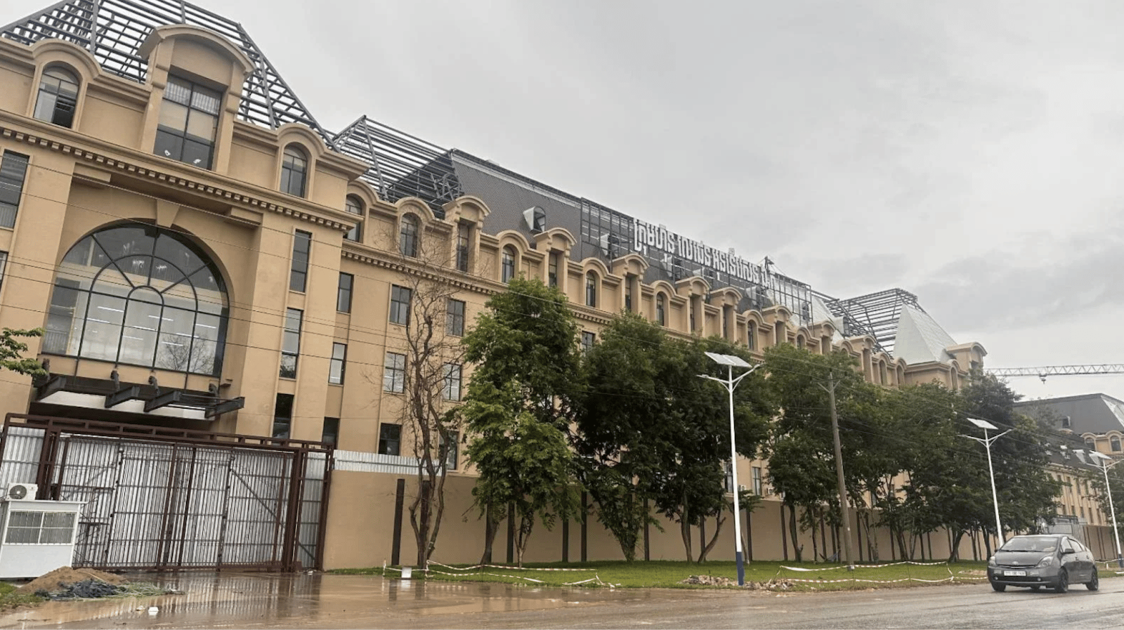 Large building with a classical architectural style, featuring multiple windows, trees in front, and a wet ground under cloudy skies.