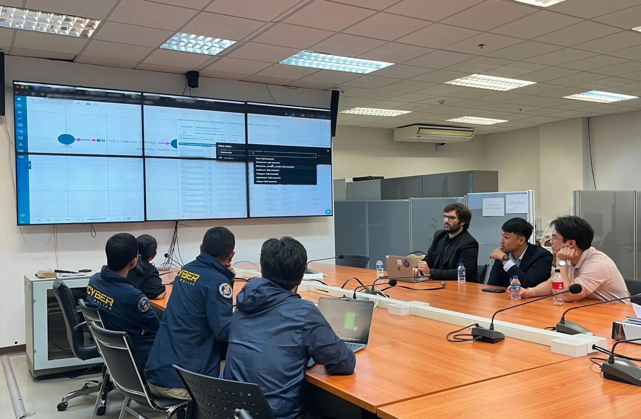 A group of individuals in a conference room watching a presentation on a large screen, with laptops and equipment visible on the table.