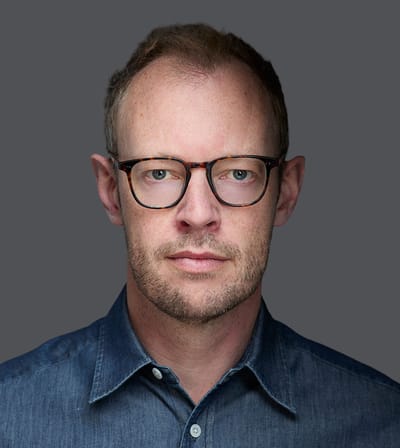 Person with short hair, wearing glasses and a denim shirt, looking directly at the camera with a neutral expression on a gray background.