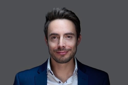 Smiling person in a dark suit with a white shirt, posing against a neutral background. The expression conveys confidence and approachability.