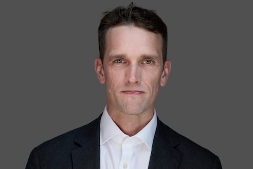 Individual with short brown hair, wearing a white shirt and dark suit jacket, facing the camera with a neutral expression against a gray background.