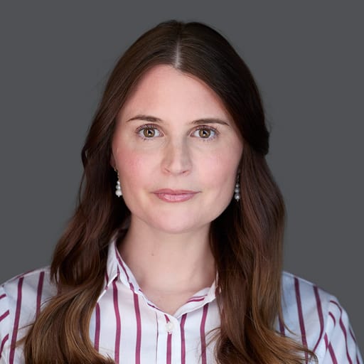 Person with long, wavy brown hair, wearing a striped button-up shirt, and earrings, posing against a plain gray background.