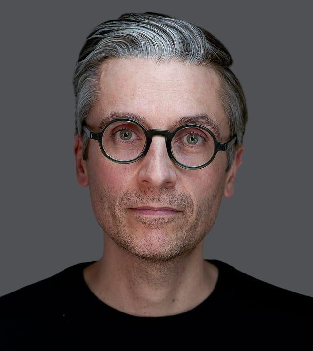 Person with gray hair and glasses, wearing a black shirt, against a gray background, looking directly at the camera.