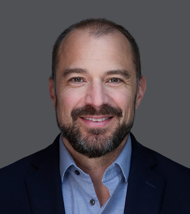 Smiling person with short hair and a beard, wearing a dark blazer and light blue shirt, set against a gray background.