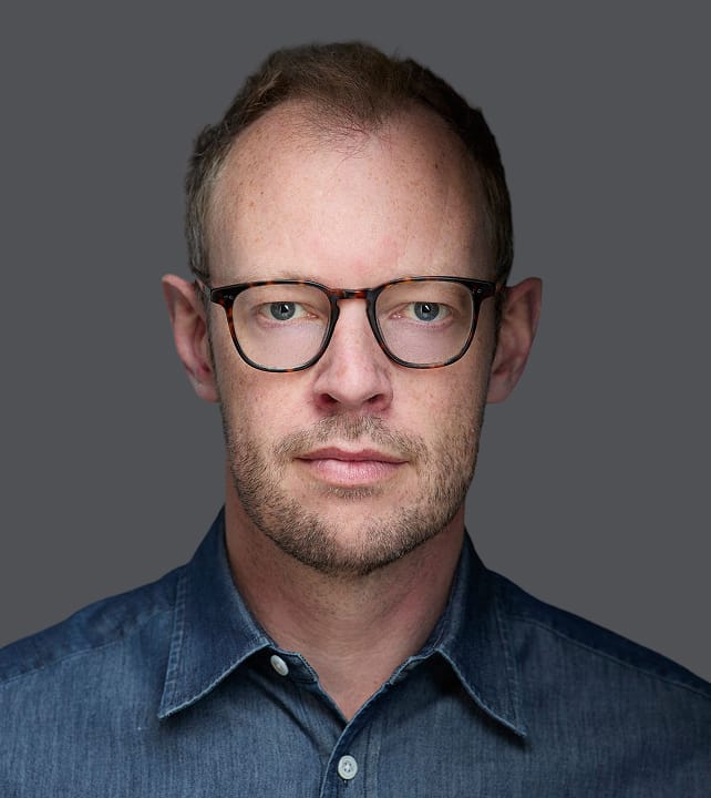 Person with short hair, wearing glasses and a denim shirt, looking directly at the camera with a neutral expression on a gray background.