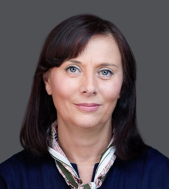 A person with shoulder-length dark hair and blue eyes, wearing a dark blouse and a patterned scarf, set against a grey background.
