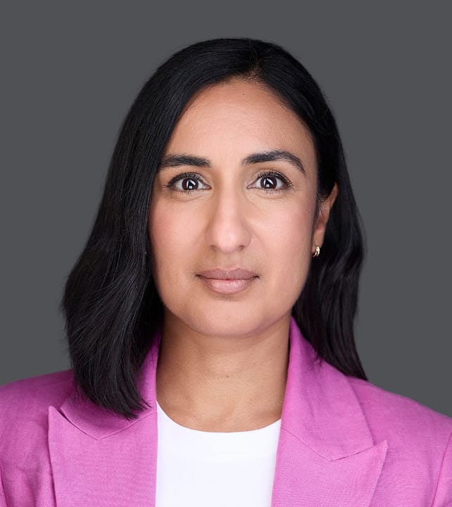 Person with dark hair wearing a light pink blazer against a neutral gray background, looking directly at the camera with a neutral expression.