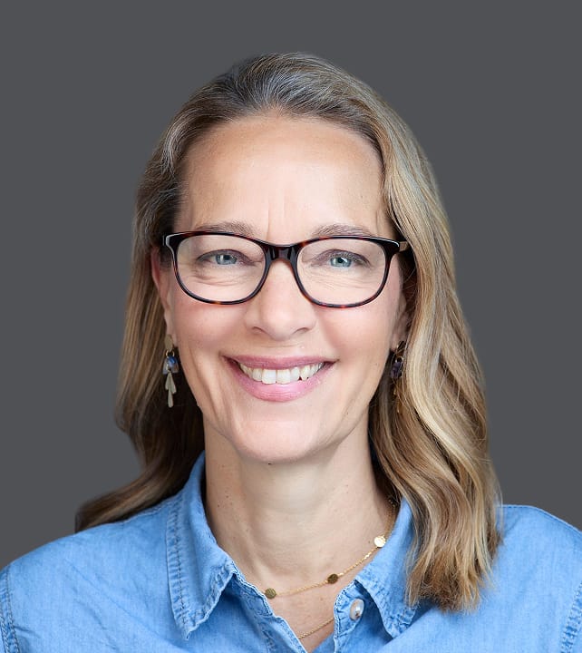 Smiling person with glasses and light brown hair wearing a denim shirt, set against a neutral gray background.
