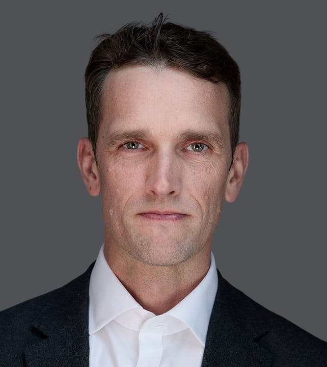 Person with short, light brown hair wearing a dark suit jacket and white shirt, looking directly at the camera against a gray background.