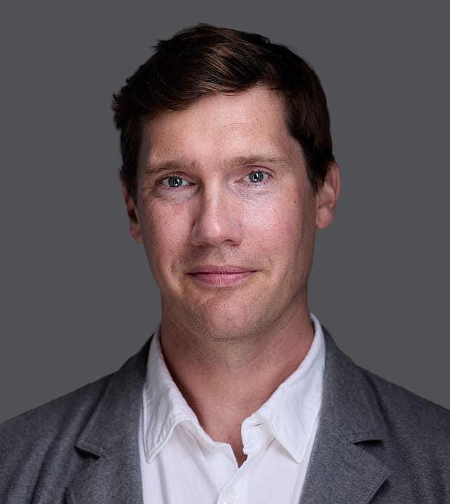 Person with short, dark hair and blue eyes, wearing a light shirt and a gray blazer, against a neutral gray background.
