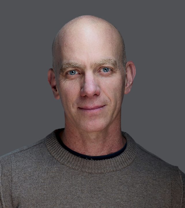 Bald individual with blue eyes wearing a gray sweater, posing against a neutral gray background.
