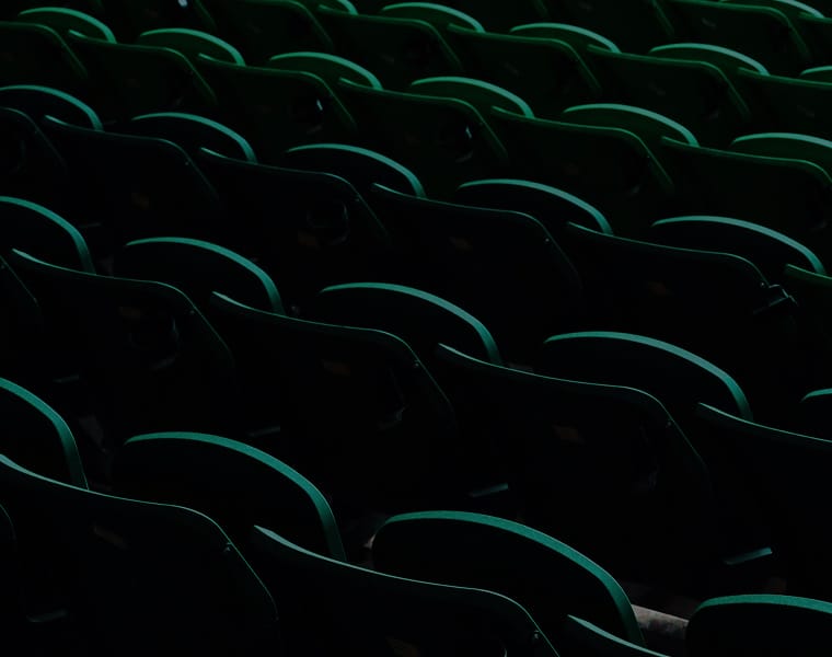Rows of dark green stadium seats arranged in a pattern, set against a dim background, creating a sense of depth and emptiness.