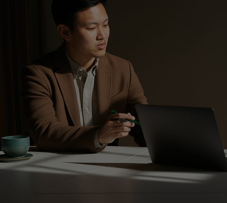 Person wearing a brown blazer and white shirt sitting at a table with a laptop and a cup, holding a green pen, in low light.