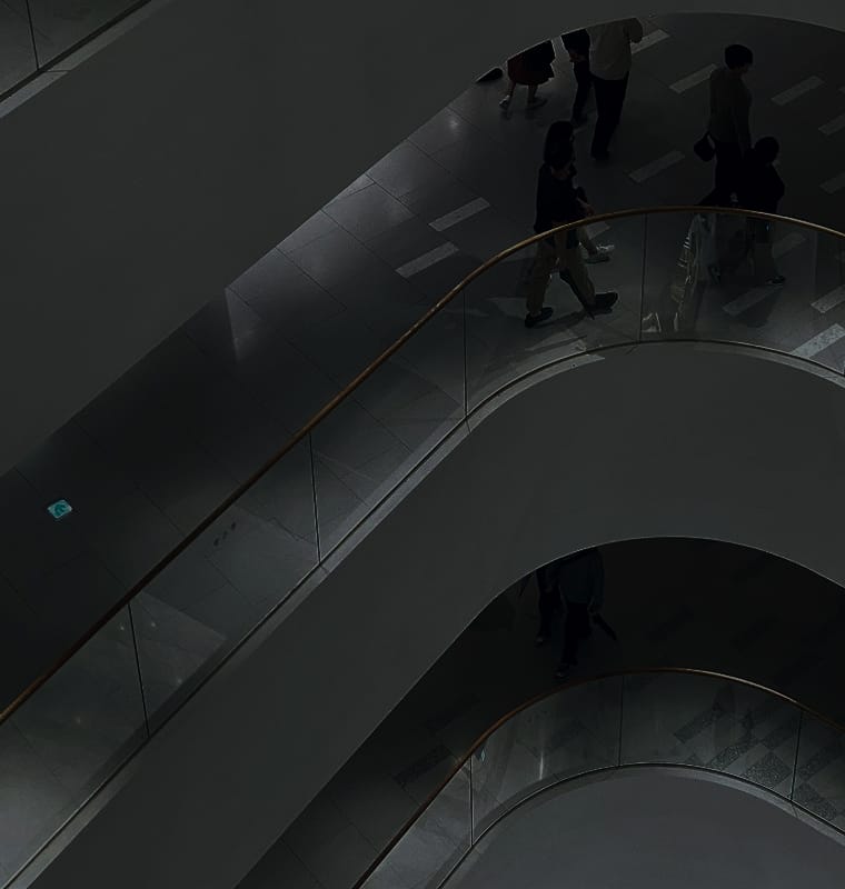 A high-angle view of a modern staircase with curved glass railings and people walking below on the tiled floor.
