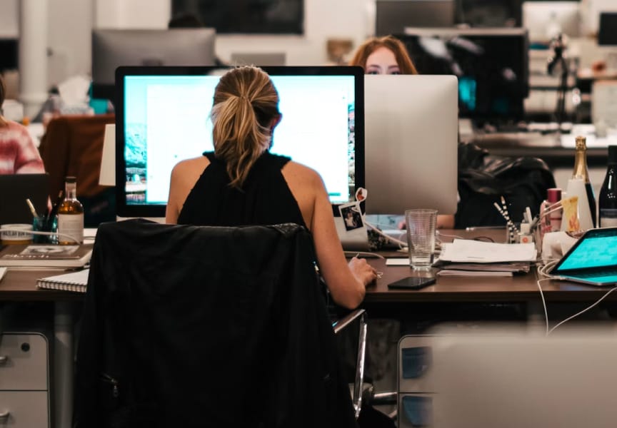 An office environment with two individuals working on computers at desks cluttered with various items, including drinks and documents.
