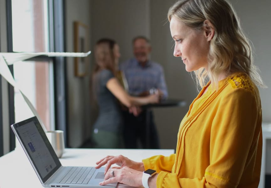 A person in a yellow blouse working on a laptop at a table, with two people engaging in conversation in the background.